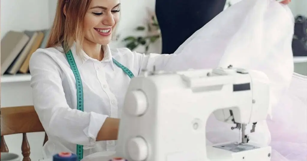 Mujer joven y sonriente trabajando desde su casa con una máquina de coser moderna confeccionando una prenda de tela blanca.