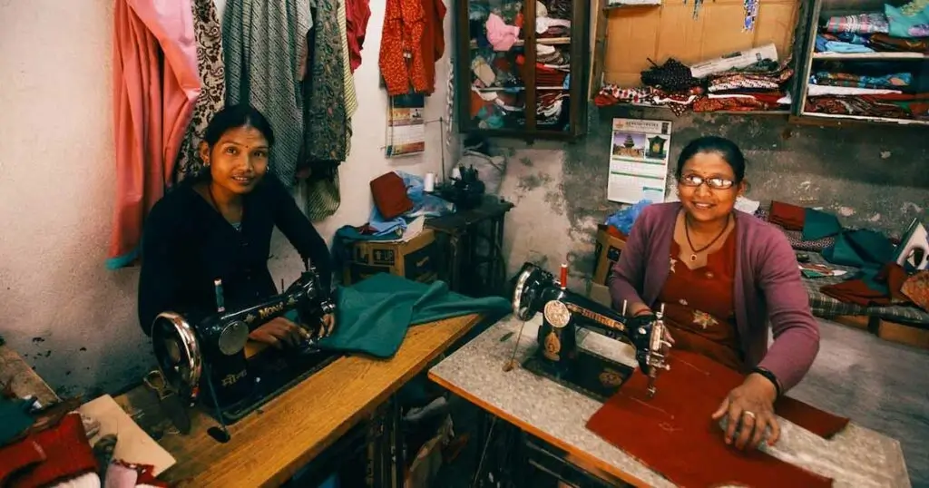 Dos mujeres costureras sonriendo mientras trabajan con máquinas de coser clásicas de color negro en un pequeño taller de confección textil.