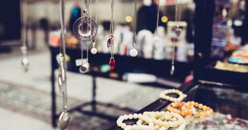 Muestra de collares de plata con diversos colgantes y dijes suspendidos en un expositor de tienda, con pulseras desenfocadas en primer plano.
