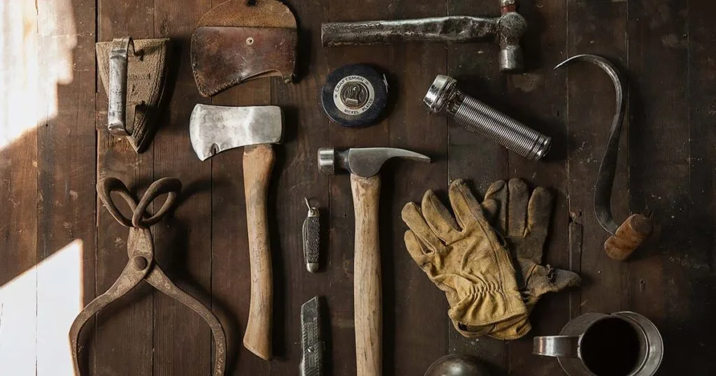 Vista superior de herramientas de trabajo rústicas sobre una mesa de madera oscura, incluyendo un martillo, hacha, guantes de cuero, pinzas y linterna para labores de ensamblaje.