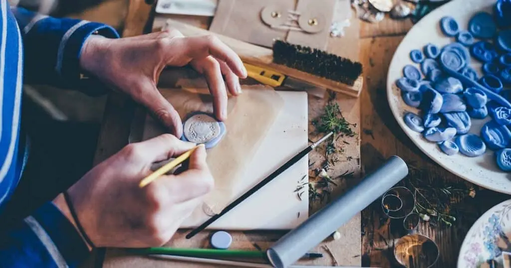 Manos de un artista modelando pequeñas piezas de arcilla o masa azul utilizando herramientas de escultura y detalle sobre una superficie de trabajo de madera.