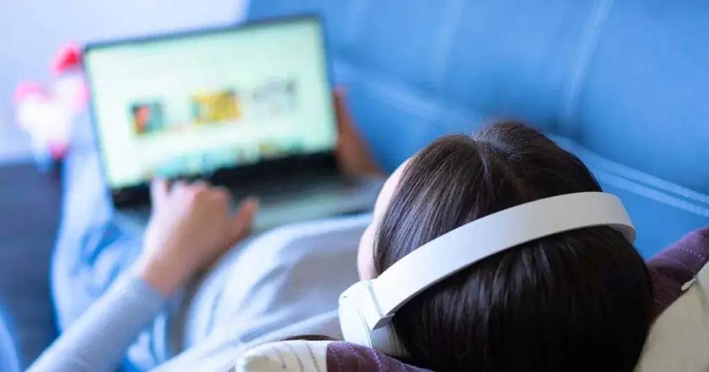 Mujer joven con auriculares recostada, trabajando cómodamente desde casa en su laptop como digitador remoto.