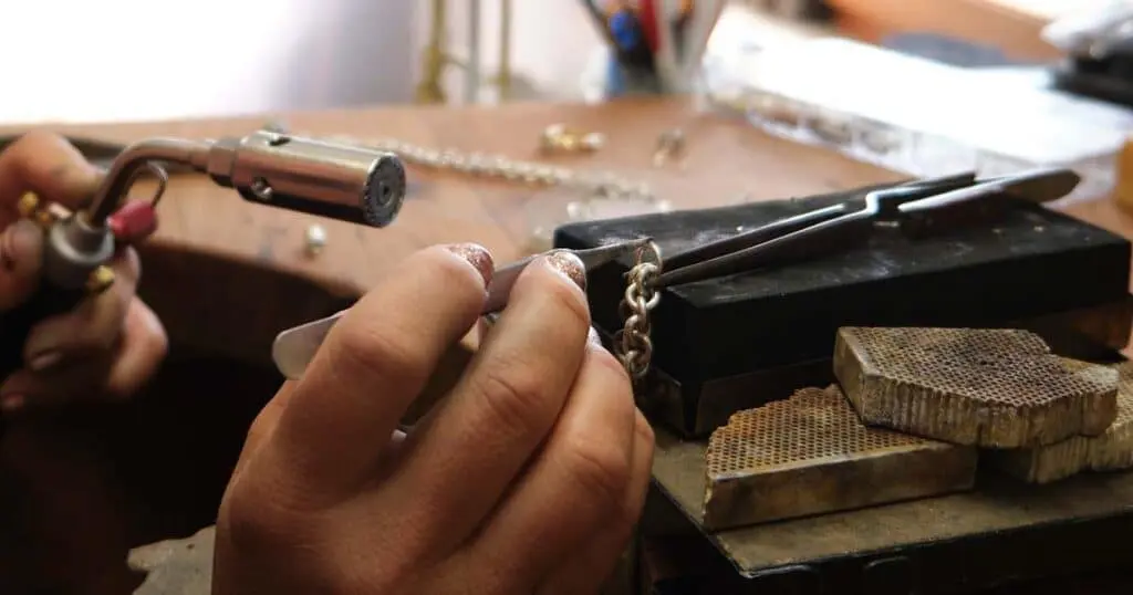 Primer plano de las manos de un joyero utilizando un mini soplete y pinzas de precisión para soldar los eslabones de una cadena de plata sobre un bloque refractario.